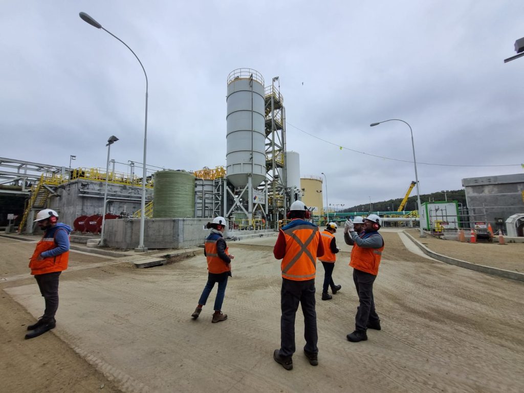 Fotografía de unas pocas personas con chalecos reflectantes naranjos y casos blancos mirando hacia distintas direcciones en las afueras de una planta desaladora
